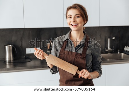 Picture of young beautiful woman standing in kitchen holding rolling pin in hands. Looking at camera.