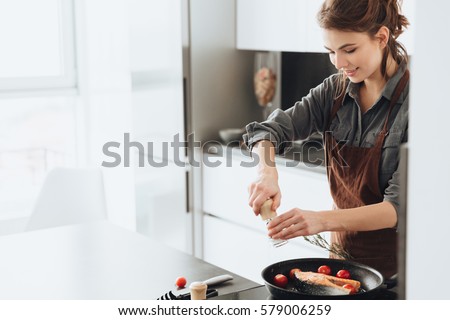 Photo of young pretty lady standing in kitchen while cooking fish. Looking aside. Royalty-Free Stock Photo #579006259