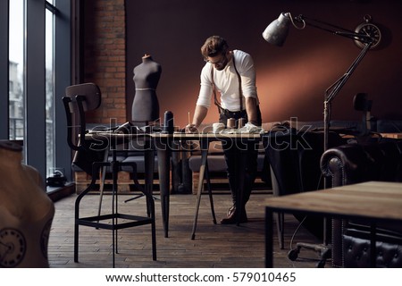 Handsome serious tailor in white shirt with brown leather suspenders working near wooden table with threads, apron and scissors in amazing atelier with antique furniture and mannequin on background Royalty-Free Stock Photo #579010465