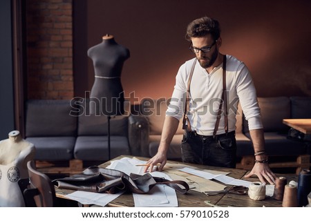 Cute tailor male with beard and glasses in white shirt with brown leather suspenders working near wooden table with threads, apron and scissors in amazing atelier with antique furniture  Royalty-Free Stock Photo #579010528