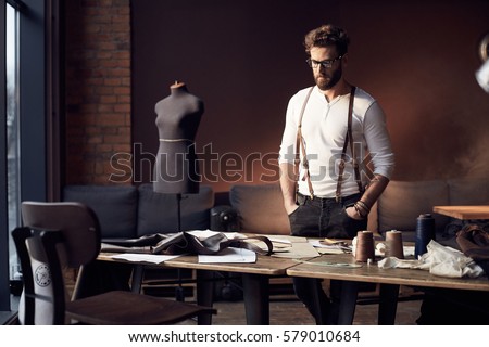 Serious tailor in white shirt with brown leather suspenders standing near wooden table with threads and tailoring equipment in amazing atelier with antique furniture and mannequin on background Royalty-Free Stock Photo #579010684