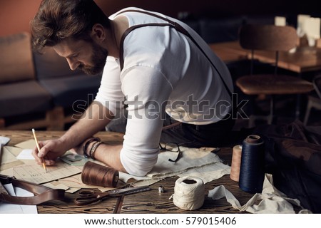 Handsome tailor male with beard in white shirt with brown leather suspenders working near wooden table with threads in amazing atelier with antique furniture and mannequin on background Royalty-Free Stock Photo #579015406