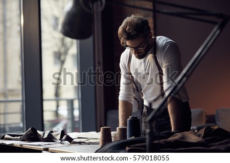 Young serious tailor with beard and glasses in white shirt with leather suspenders looking on drawing near wooden table with threads in amazing atelier with antique furniture and mannequin  Royalty-Free Stock Photo #579018055