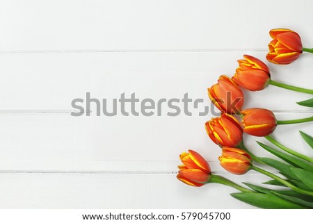 Overhead shot a bouquet of orange and yellow Spring tulip flowers and blank paper card for Mother's or Women's Day over white wood table top. Flat lay top view style.