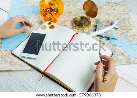 Bloger planning budget of trip Hands holding pen near notebook Table with traveling theme items mobile phone, map, compass, plane and piggy bank Empty mockup notes, your can place text logo images.  Royalty-Free Stock Photo #579066373