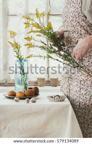 Preparing for Easter. Female hands cutting by old scissors yellow flowers near white tablecloth table decorated brown chicken and quail colored easter eggswith window as background. Day light. Toned