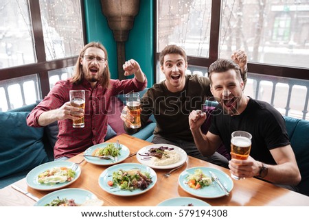 Picture of cheerful men friends sitting in cafe while eating and drinking beer.
