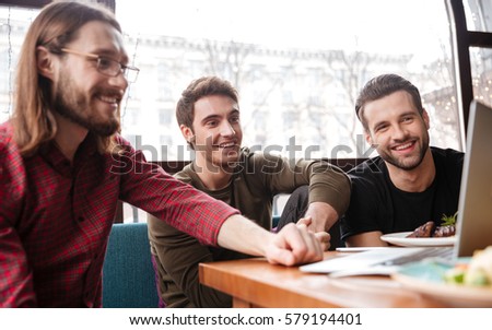 Picture of cheerful men friends sitting in cafe while eating. Using laptop.