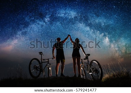 Night starry sky over cyclists on the hill. Guy and the girl with mountain bikes keep the hands lifted upwards to sky. Night landscape with colorful Milky Way. Bottom view