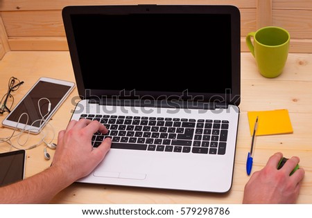 Laptop on a table. A man is typing on the keyboard, next to the green computer mouse, green cup, headphones, smart phone, tablet.