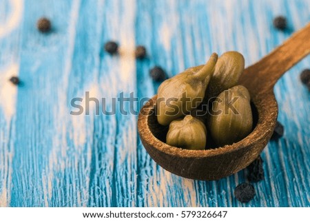 Horizontal photo with several green capers which are placed in vintage wooden spoon. Few pieces of whole pepper seeds spice are spilled around. All is placed on blue wooden board.
