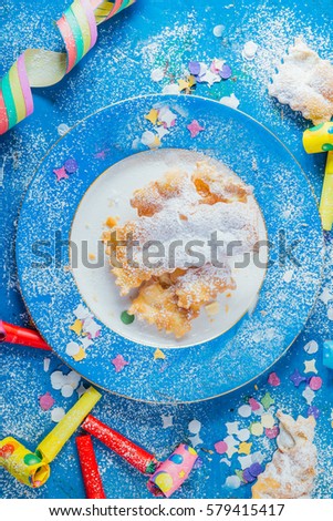 Carnivalesque composition. Top view of an elegant blue dish with sfrappols or chiacchere,italian fried carnival cookies with icing sugar , around whistles, confetti on a bluish shabby chic board.