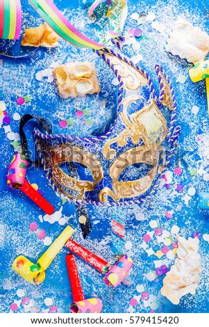 Carnivalesque composition. Top view of a carnival mask, whistles, confetti, and sfrappoles or chiacchere,italian fried carnival cookies with icing sugar on a bluish shabby chic board.