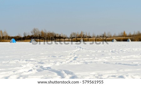 Small tents for winter fishing standing on a frozen lake in the snow next to the old dry grass. 