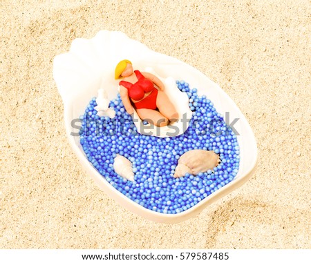 Fat blonde doll in shells on sand. Handmade model of female tourist on white sand beach. Artificial sea water made of blue balls. Sunny day by the seaside. Small puppet macro photo with sand texture