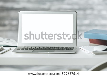 Close up of empty white laptop screen, books and other items on light desktop. Blurry wooden background. Mock up