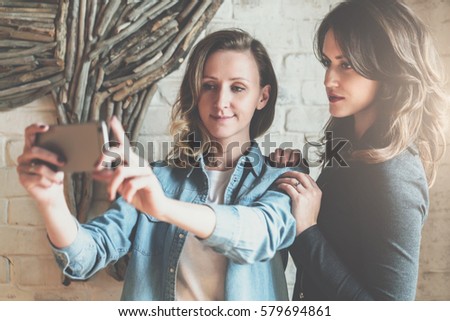 One-on-one meeting. Two young businesswomen doing selfie in restaurant. In background white brick wall. Meeting friends, dinner together. Girls use digital gadgets. Girls are photographed in cafe.