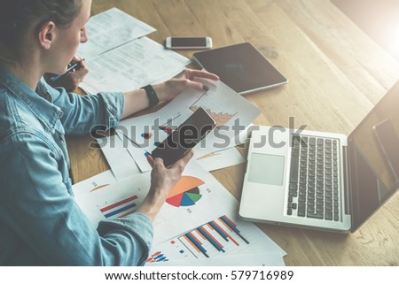 View from above. Young business woman sitting in office at desk, looking at graphics and uses smartphone. On table is laptop and tablet computer. Nearby sits other girl and sign papers. Teamwork.