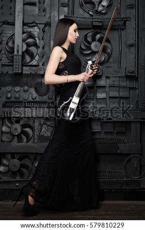 A slender woman in a black dress standing with a violin sideways to the photographer. On the background of the black metal door. Vertical photo
