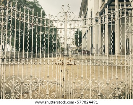 Details of a white and aged arabesque closed gate that leads to a church courtyard with ground floor and trees on a cloudy cold day