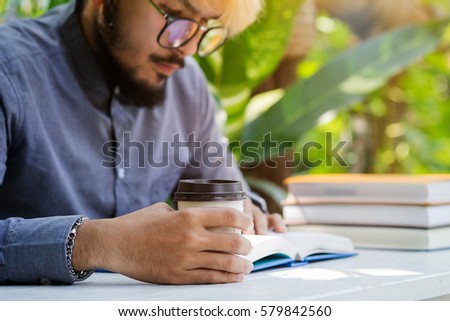 Man sitting on a bench holding a cup of coffee outside and read a book.The young students sitting the exam preparation book.Focus on hands