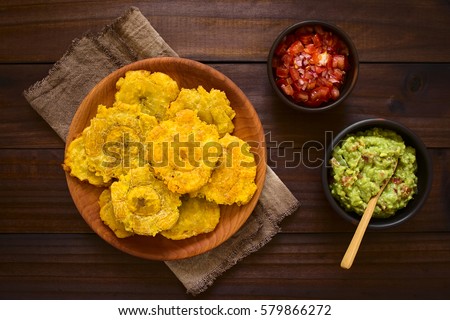 Patacon or toston fried and flattened pieces of green plantain, traditional snack or accompaniment in the Caribbean, guacamole and tomato onion salad beside, photographed overhead with natural light Royalty-Free Stock Photo #579866272