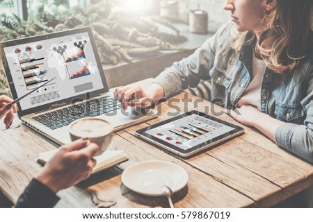 Teamwork,two young business women sitting at table,drinking coffee and working online.First girl is showing pen on chart and diagram on computer screen.On table tablet PC.Students are learning online. Royalty-Free Stock Photo #579867019