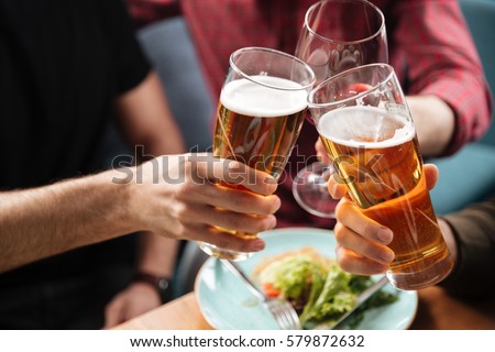 Cropped photo of young friends sitting in cafe while drinking alcohol. Focus on glasses of beer. Royalty-Free Stock Photo #579872632