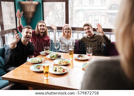 Photo of young happy friends sitting in cafe while waving to friend.