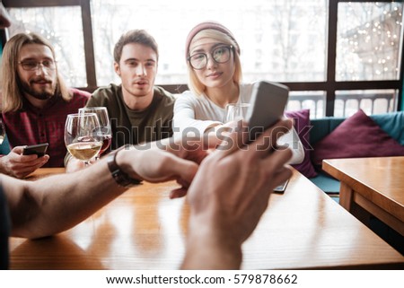 Image of smiling friends sitting in cafe and drinking alcohol and looking at phone.