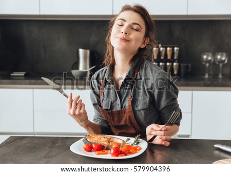 Image of pretty young woman sitting in kitchen while eating fish and tomatoes. Royalty-Free Stock Photo #579904396