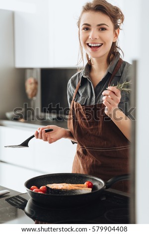 Picture of young happy woman standing in kitchen while cooking fish. Looking at camera.