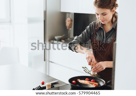 Picture of young happy lady standing in kitchen while cooking fish. Looking aside.