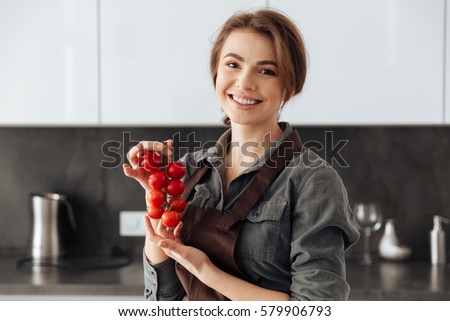 Picture of young happy woman standing in kitchen holding tomatoes in hands.
