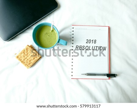 a notebook with text 2018 resolution, cup of green tea, biscuit and laptop over white background