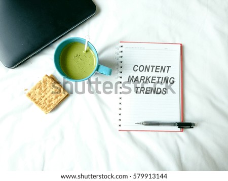 a notebook with text content marketing trends, cup of green tea, biscuit and laptop over white background