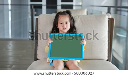 Cute little child girl holding empty green blackboard on fabric sofa in library room.
