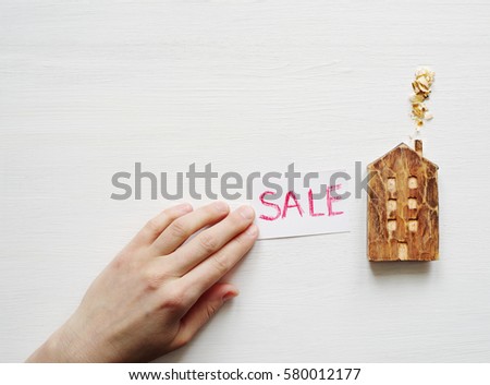 Wooden house and a white card with the red inscription "SALE" on a white wooden background. Conceptual image of sale of the house
