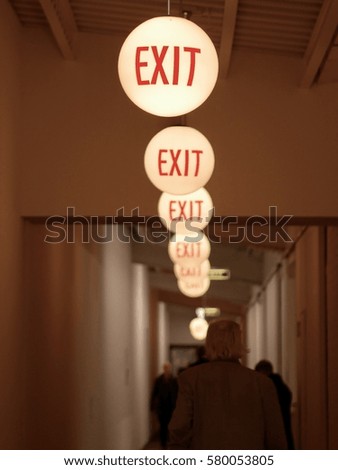 Row of round illuminated ceiling lights in a corridor or passage with walking people marking the route to the exit with red text as a safety measure in case of emergencies