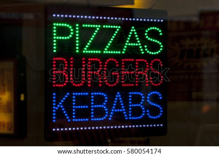 A neon sign in a takeaway shop window advertising Pizzas, Burgers, and Kebabs.