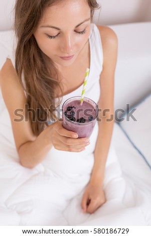 Beautiful girl sitting on a bed after waking up in the morning, smiling and holding a glass of raspberry and blueberry mix smoothie and smiling