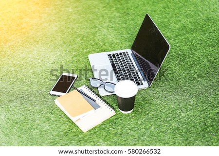 Office on the grass. On the lawn there is a laptop with a blank screen, next to it is a cup of coffee, black notebook, pen, smart phone and wired headphones.Mock up.Gadgets on the grass.