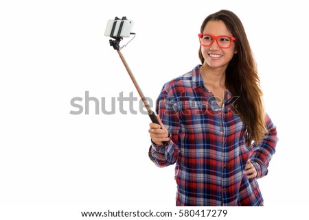 Studio shot of young happy woman smiling while holding selfie stick and taking selfie picture with mobile phone