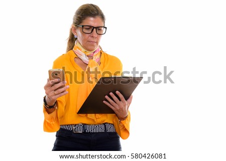 Studio shot of mature beautiful businesswoman holding mobile phone while reading on clipboard