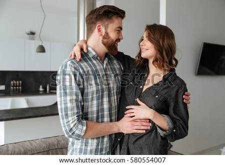 Photo of young happy loving couple hugging in kitchen at home indoors. Looking at each other.