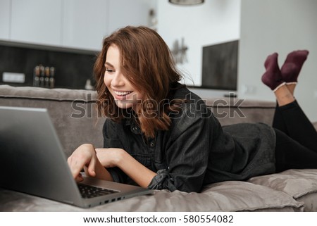 Photo of young cheerful lady in kitchen at home indoors lies on sofa and using laptop computer.