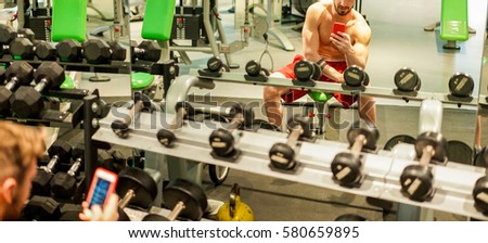 Young fitness personal trainer using mobile smartphone inside gym - Athlete having break after workout training - Healthy lifestyle and bodybuild concept - Focus on him glass reflection - Warm filter