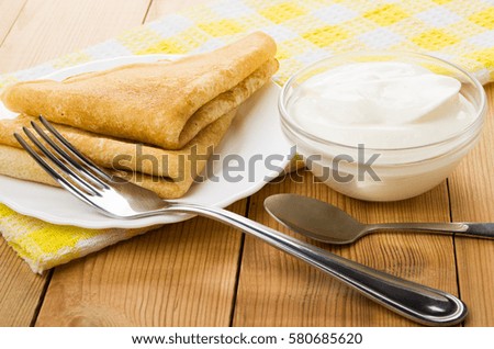 Folded russian pancakes in plate, fork and bowl with sour cream on wooden table