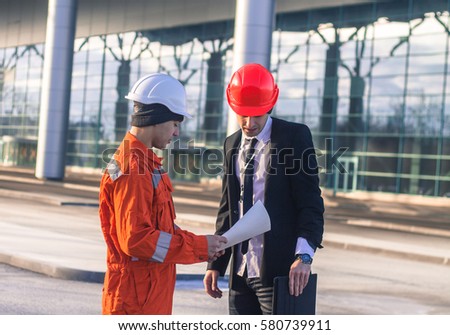 young boss and worker in conversation discussing a construction project. They wear  safety helmets. Tablet in hand. Business modern background
