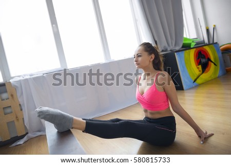 Young beautiful girl in a pink shirt and black tights to perform acrobatic workout on the mat for yoga in the gym.